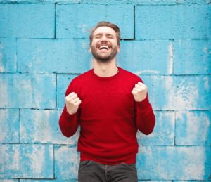 A man wearing a red sweater stands in front of a blue brick wall smiling with clenched fists, feeling successful