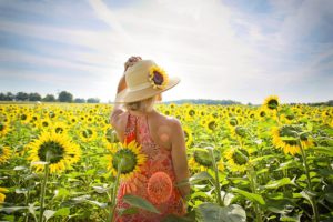 A woman holding onto her sun hat stands facing a field of sunflowers