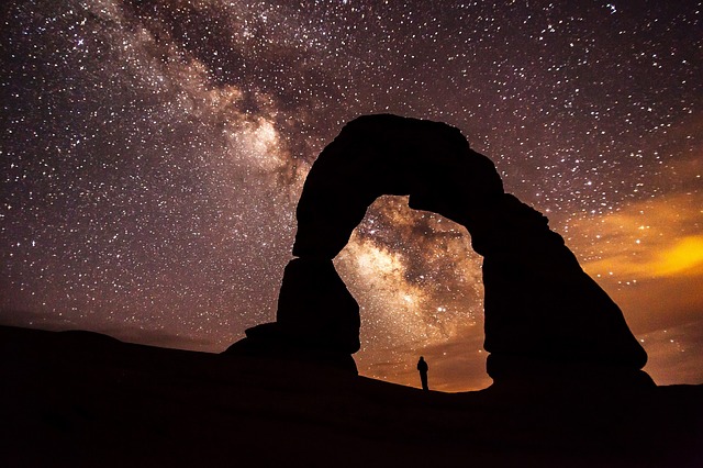 A figure stands under a giant rock arch under a stary sky