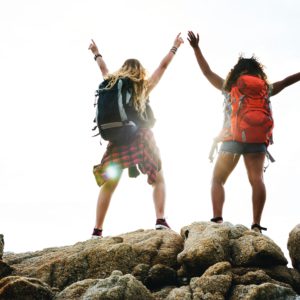Two women wearing backpacks stand with arms above their heads successfully climbed a mountain